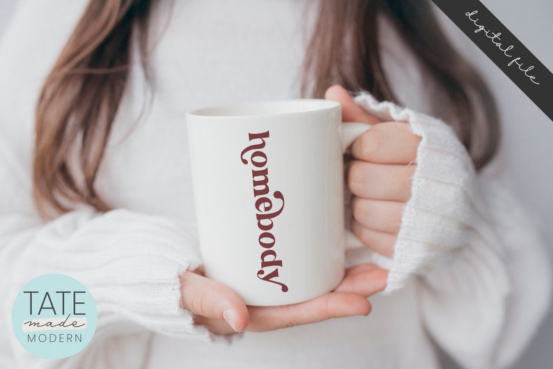 May include: White ceramic mug with the text "homebody" written vertically in a maroon color.