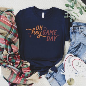 May include: A navy blue t-shirt with the text "OH hey GAME DAY" in orange and red lettering. The shirt is laid out on a white surface with a plaid blanket, blue jeans, and white sneakers.