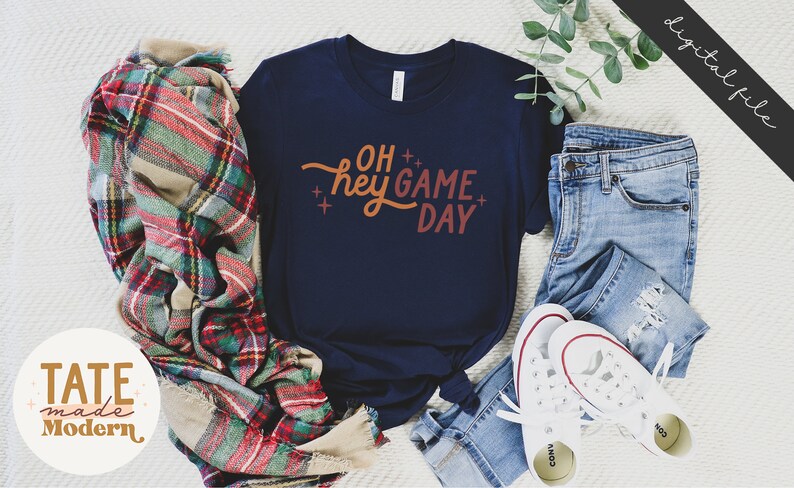 May include: A navy blue t-shirt with the text "OH hey GAME DAY" in orange and red lettering. The shirt is laid out on a white surface with a plaid blanket, blue jeans, and white sneakers.
