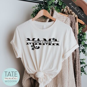 Op de afbeelding: Een wit T-shirt met een zwarte afbeelding waarop "Mama" staat, met daaronder een lijn met de tekst "Emery + Rylan" en bloemmotieven.