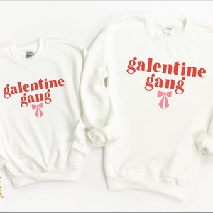 May include: Two white crewneck sweatshirts with the text "galentine gang" in red letters and a pink bow.