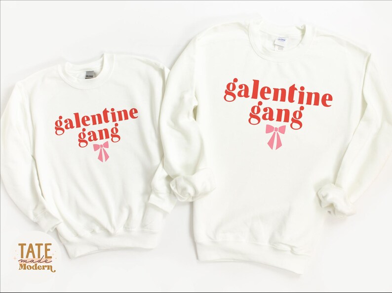 May include: Two white crewneck sweatshirts with the text "galentine gang" in red letters and a pink bow.