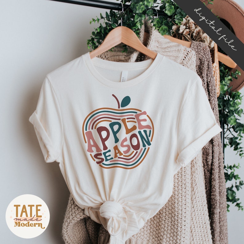 May include: A white t-shirt with a graphic of an apple with the text "APPLE SEASON" inside the apple. The apple is made of colorful stripes in shades of pink, orange, brown, and teal.