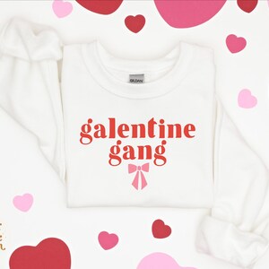 May include: White sweatshirt with red text that says "galentine gang" and a pink bow. The sweatshirt is surrounded by pink and red hearts.