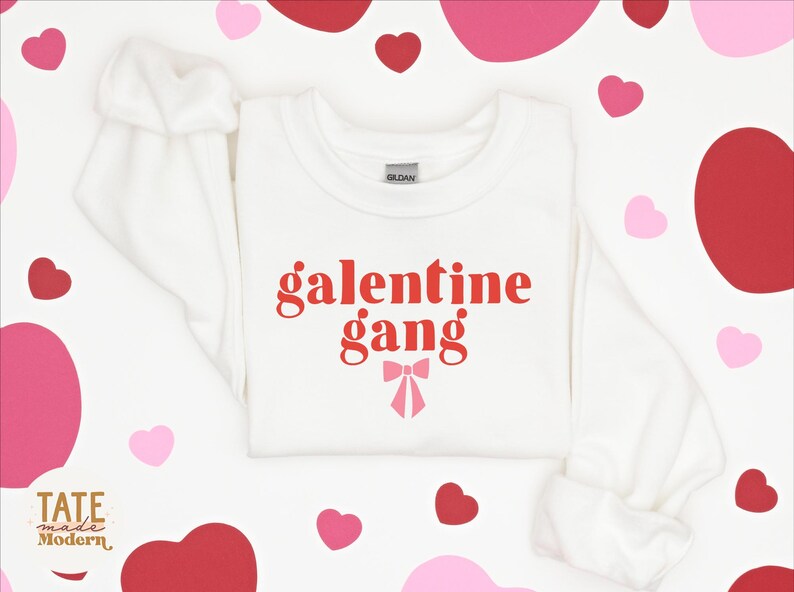 May include: White sweatshirt with red text that says "galentine gang" and a pink bow. The sweatshirt is surrounded by pink and red hearts.