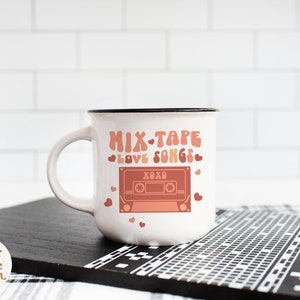 May include: White ceramic mug with a black rim featuring a pink and red cassette tape graphic with the text "Mix Tape Love Songs" and hearts.