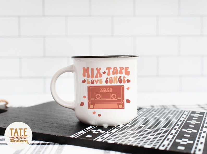 May include: White ceramic mug with a black rim featuring a pink and red cassette tape graphic with the text "Mix Tape Love Songs" and hearts.