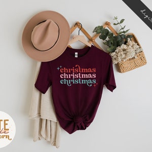 Pode incluir: Uma camiseta vinho com o texto "christmas christmas christmas" em uma fonte retr&ocirc; com cores diferentes. A camiseta est&aacute; pendurada em um cabide com um chap&eacute;u e uma planta em uma cesta atr&aacute;s.