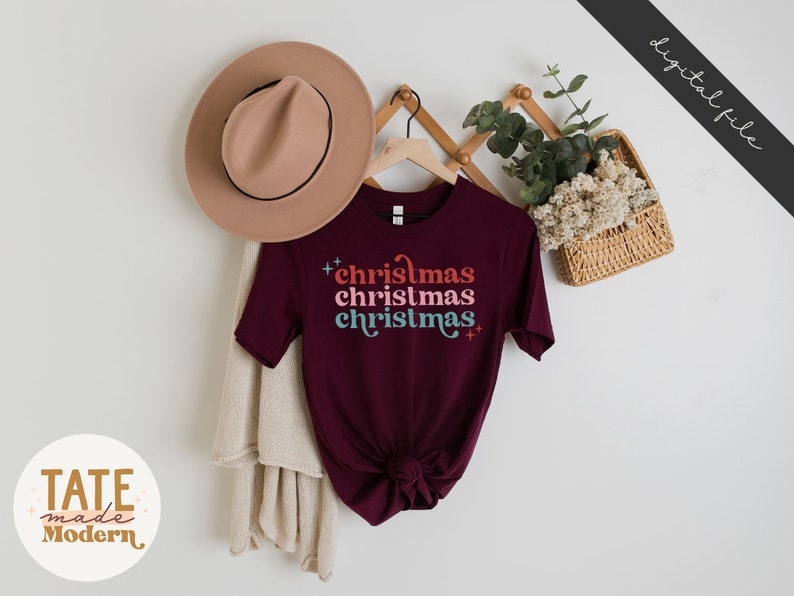 Pode incluir: Uma camiseta vinho com o texto "christmas christmas christmas" em uma fonte retr&ocirc; com cores diferentes. A camiseta est&aacute; pendurada em um cabide com um chap&eacute;u e uma planta em uma cesta atr&aacute;s.