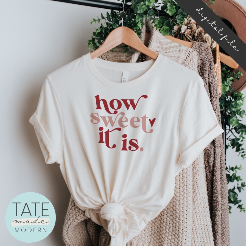 May include: A white t-shirt with the text "how sweet it is." in red and pink lettering. The shirt is on a hanger and folded in half.