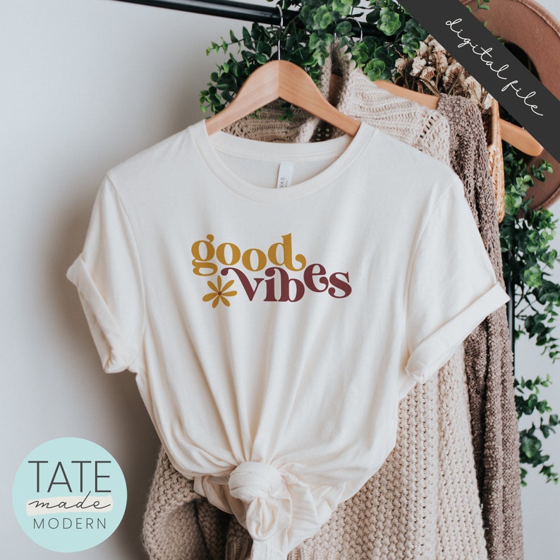 May include: A white t-shirt with the text "good vibes" in yellow and brown lettering with a small flower graphic. The shirt is on a hanger and folded.