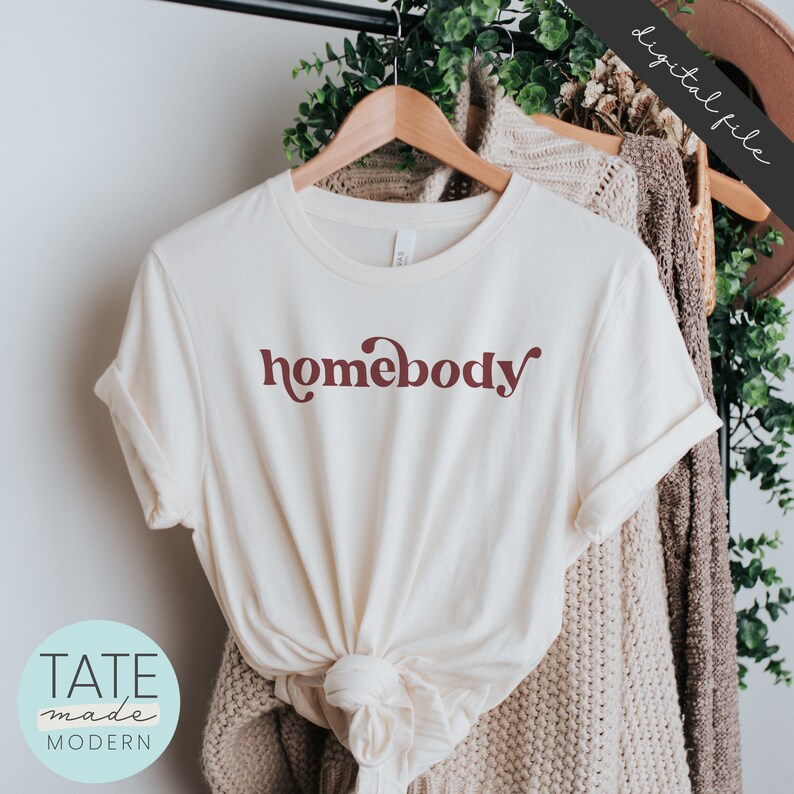 May include: A white t-shirt with a maroon "homebody" graphic in a retro font style. The shirt is tied in a knot at the bottom and is hanging on a wooden hanger.