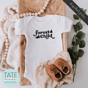 May include: White baby onesie with black text that reads "forest child" and a graphic of stylized trees. The onesie is laid out on a wooden surface with a brown blanket and brown shoes.