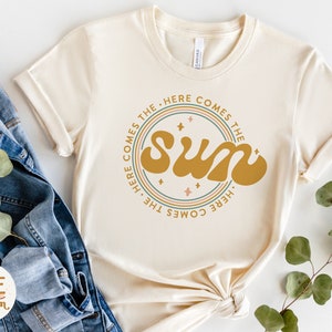 Pu&ograve; includere: Una maglietta beige con un design grafico circolare che presenta il testo "Here comes the sun" in un font retr&ograve;. Il grafico &egrave; circondato da tre cerchi concentrici in tonalit&agrave; di rosa, verde e blu.