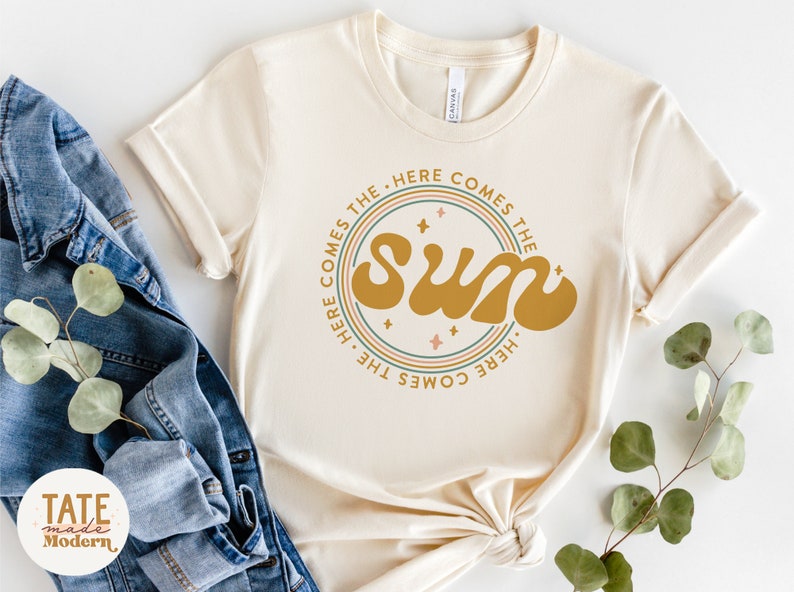 Pu&ograve; includere: Una maglietta beige con un design grafico circolare che presenta il testo "Here comes the sun" in un font retr&ograve;. Il grafico &egrave; circondato da tre cerchi concentrici in tonalit&agrave; di rosa, verde e blu.