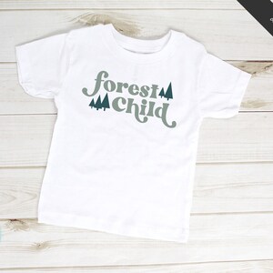 May include: White t-shirt with the text "forest child" in a retro font with green trees on either side of the text.