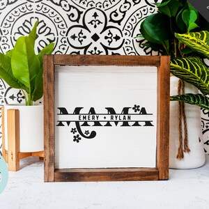 Op de afbeelding: Een houten ingelijst bord met het woord "MAMA" in zwarte letters met een decoratief bloemenmotief. Het bord bevat ook de namen "EMERY + RYLAN" in een kleiner lettertype.