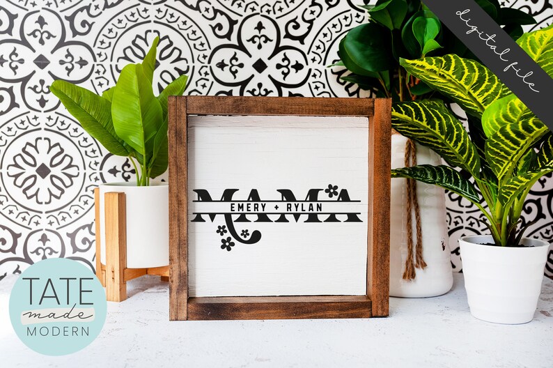 Op de afbeelding: Een houten ingelijst bord met het woord "MAMA" in zwarte letters met een decoratief bloemenmotief. Het bord bevat ook de namen "EMERY + RYLAN" in een kleiner lettertype.
