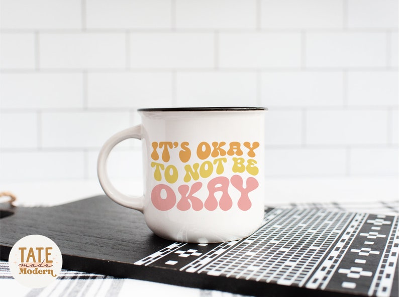 Puede incluir: Taza de cer&aacute;mica blanca con borde y asa negros. La taza tiene un gr&aacute;fico colorido que dice "It's okay to not be okay" en una fuente retro.