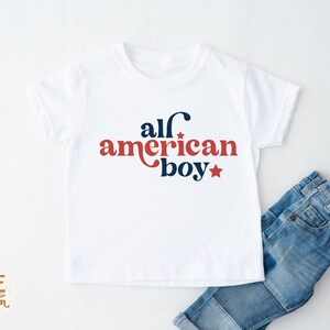 May include: White t-shirt with red and blue text that says "all american boy" with a star on either side of the text. The shirt is on a white surface with a pair of blue jeans folded on the bottom right.
