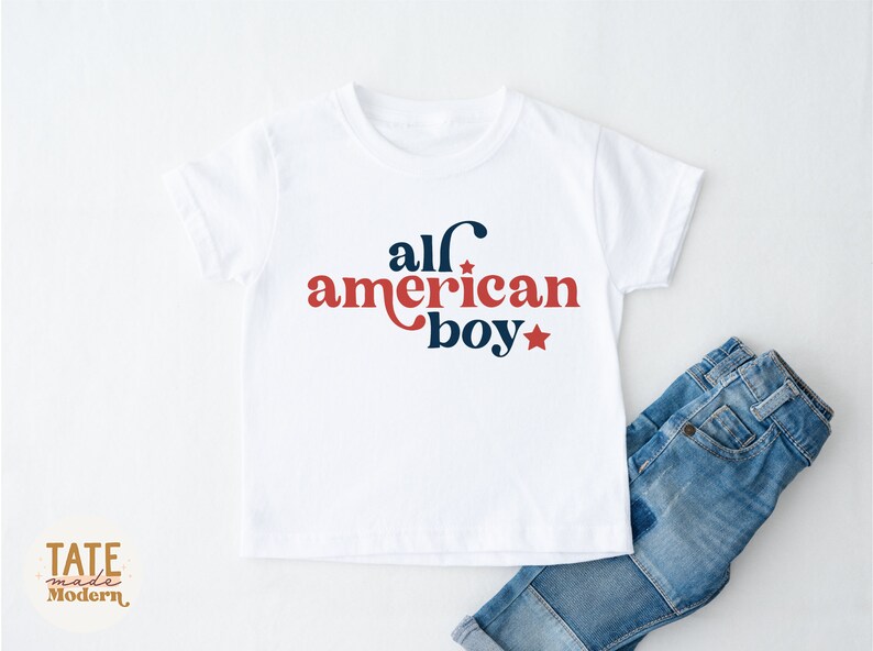 May include: White t-shirt with red and blue text that says "all american boy" with a star on either side of the text. The shirt is on a white surface with a pair of blue jeans folded on the bottom right.