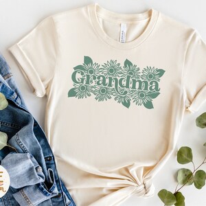 May include: A cream colored t-shirt with a green graphic of the word "Grandma" surrounded by stylized flowers.