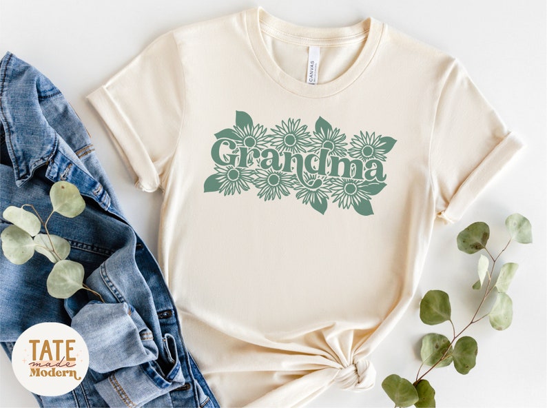 May include: A cream colored t-shirt with a green graphic of the word "Grandma" surrounded by stylized flowers.