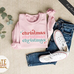 Pode incluir: Camisola cor-de-rosa com o texto "christmas christmas christmas" numa fonte retro com cores vermelho, rosa e azul.
