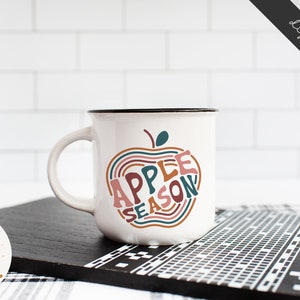 May include: White ceramic mug with a black rim and handle. The mug features a graphic of an apple with the text "Apple Season" inside the apple. The text is in a colorful, retro font.