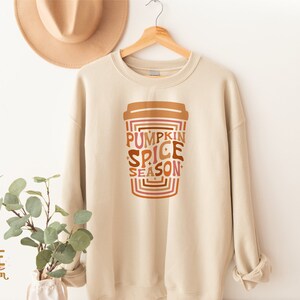 Może przedstawiać: Beżowa bluza z nadrukiem kubka z kawą, na kt&oacute;rym widnieje napis "Pumpkin Spice Season" w retro czcionce. Tekst jest w odcieniach brązu, pomarańczy i r&oacute;żu.