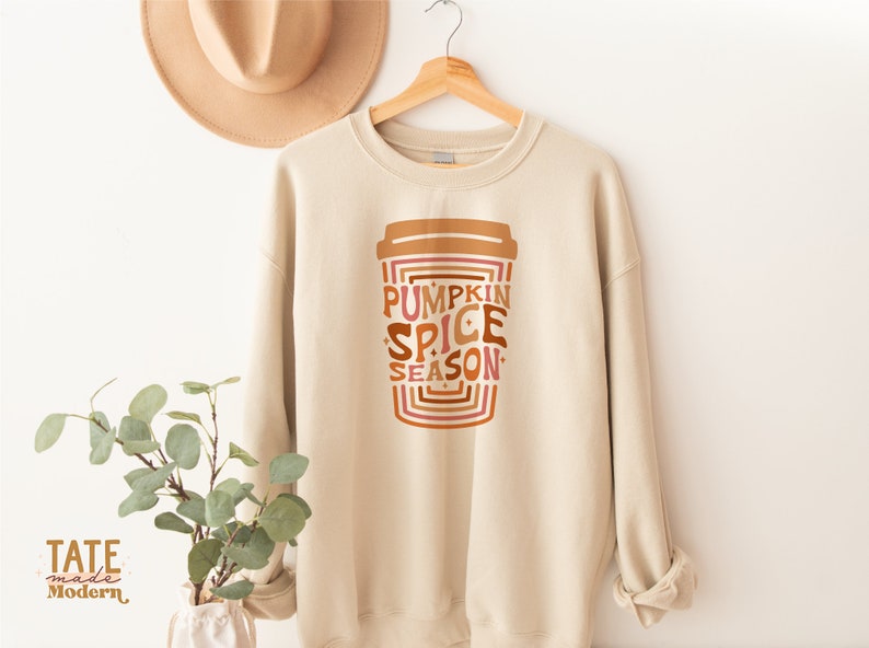 Może przedstawiać: Beżowa bluza z nadrukiem kubka z kawą, na kt&oacute;rym widnieje napis "Pumpkin Spice Season" w retro czcionce. Tekst jest w odcieniach brązu, pomarańczy i r&oacute;żu.