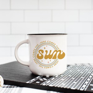 Pu&ograve; includere: Tazza in ceramica bianca con bordo e manico neri. La tazza presenta un design grafico di un sole giallo con il testo "Here comes the sun" in un cerchio attorno.