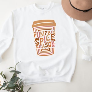 Może przedstawiać: Biała bluza z nadrukiem kubka z kawą z tekstem "Pumpkin Spice Season" w retro czcionce.