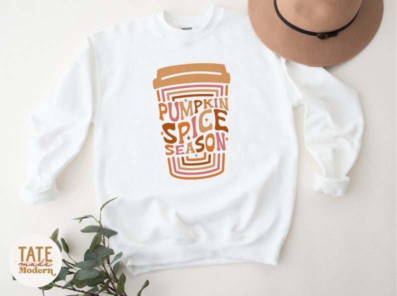 Może przedstawiać: Biała bluza z nadrukiem kubka z kawą z tekstem "Pumpkin Spice Season" w retro czcionce.
