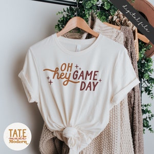 May include: A white t-shirt with the text "OH hey GAME DAY" in brown and orange lettering. The shirt is on a hanger and has a knotted bottom.