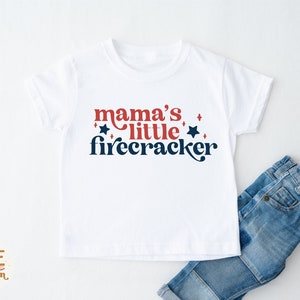 K&ouml;nnte beinhalten: Wei&szlig;es T-Shirt mit dem Text "mama's little firecracker" in Rot und Marineblau. Das Design beinhaltet Sterne und eine Schreibschrift. Blaue Jeans sind teilweise neben dem T-Shirt zu sehen.