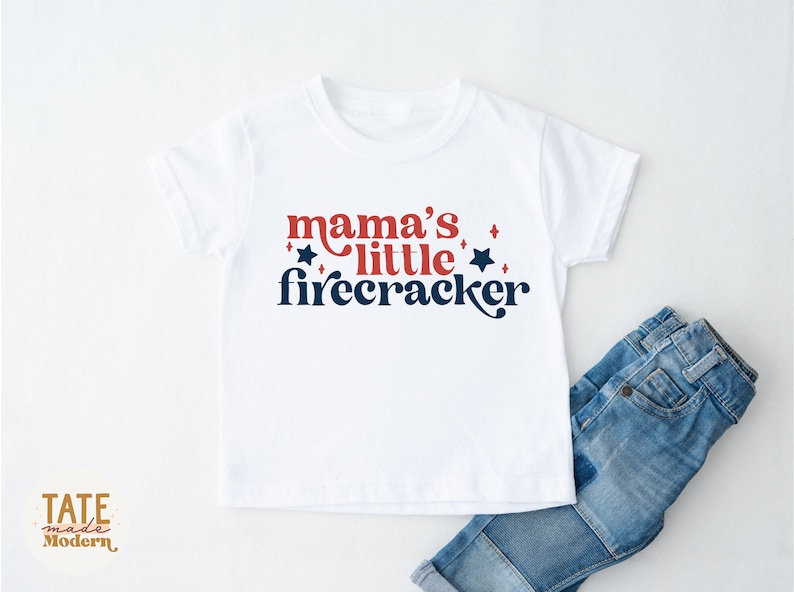 K&ouml;nnte beinhalten: Wei&szlig;es T-Shirt mit dem Text "mama's little firecracker" in Rot und Marineblau. Das Design beinhaltet Sterne und eine Schreibschrift. Blaue Jeans sind teilweise neben dem T-Shirt zu sehen.
