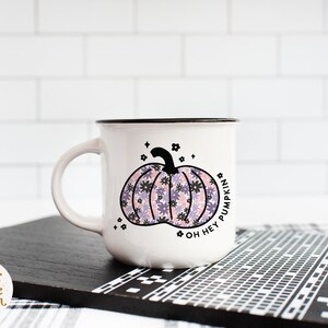 May include: White ceramic mug with a black rim and handle. The mug features a pink and purple floral pumpkin design with the text "Oh Hey Pumpkin".