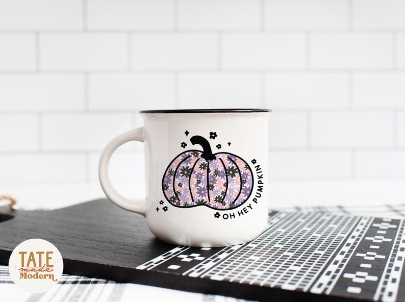May include: White ceramic mug with a black rim and handle. The mug features a pink and purple floral pumpkin design with the text "Oh Hey Pumpkin".
