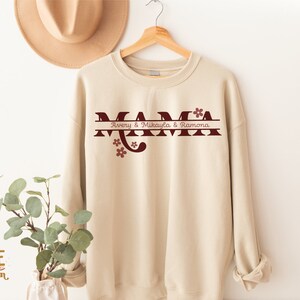 Op de afbeelding: Een beige sweatshirt met een kastanjebruine afbeelding waarop "MAMA" staat, met daaronder een gesplitste lijn met de tekst "Avery & Mikayla & Ramona". Er staan kleine kastanjebruine bloemen op de afbeelding.