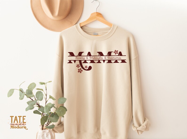 Op de afbeelding: Een beige sweatshirt met een kastanjebruine afbeelding waarop "MAMA" staat, met daaronder een gesplitste lijn met de tekst "Avery & Mikayla & Ramona". Er staan kleine kastanjebruine bloemen op de afbeelding.