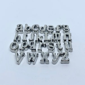 May include: A set of 26 silver metal alphabet letter charms. Each letter is individually crafted and features a slightly raised design.