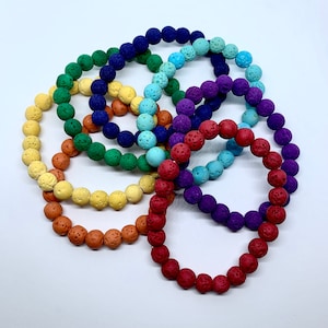 May include: A set of seven colourful lava rock bracelets. The bracelets are arranged in a rainbow pattern, with red, orange, yellow, green, blue, purple, and dark blue beads.