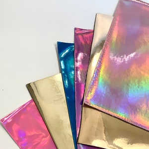 Op de afbeelding: Een verzameling van zes iriserende vinylvellen in verschillende kleuren, waaronder roze, goud, blauw en paars. De vellen zijn in een waaier vorm gerangschikt, met het bovenste vel het meest zichtbaar.
