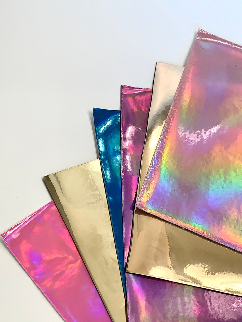Op de afbeelding: Een verzameling van zes iriserende vinylvellen in verschillende kleuren, waaronder roze, goud, blauw en paars. De vellen zijn in een waaier vorm gerangschikt, met het bovenste vel het meest zichtbaar.