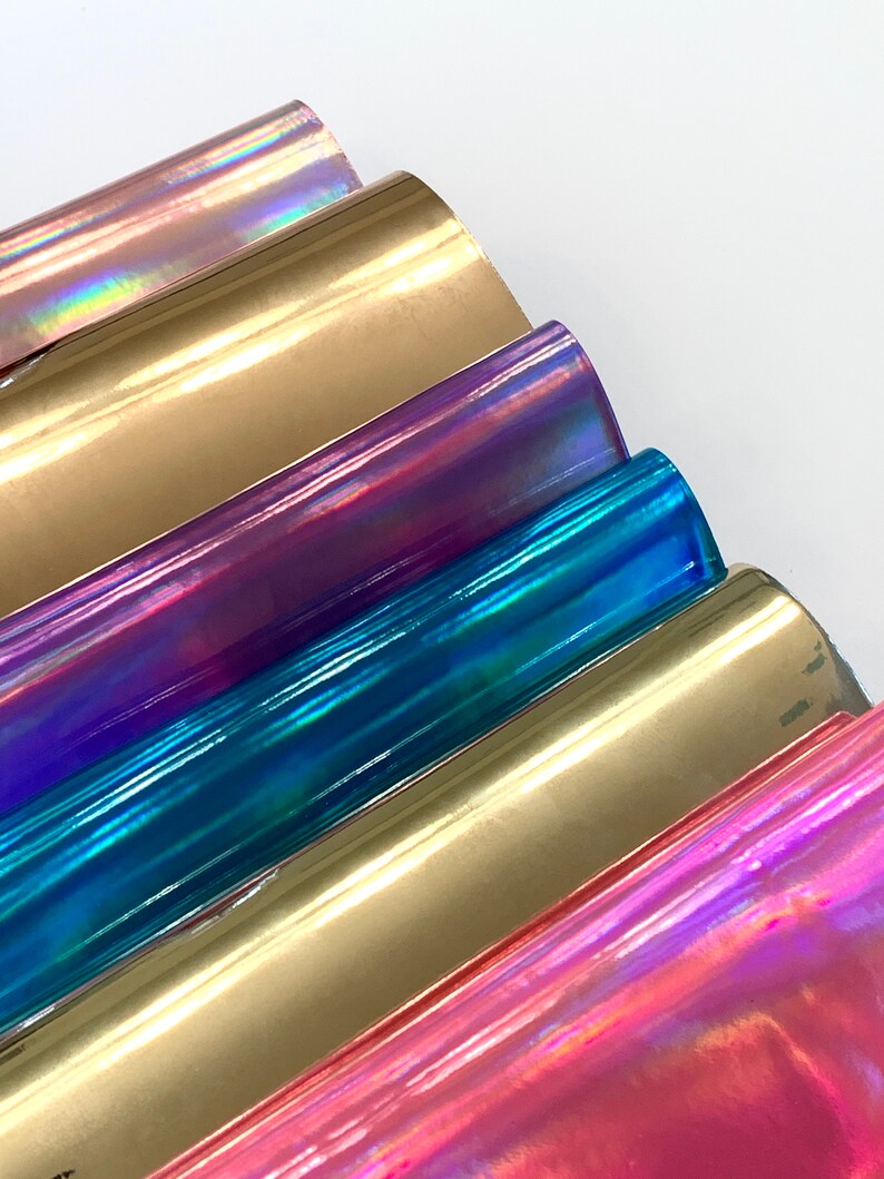 Op de afbeelding: Een set van zes iriserende vinylvellen in verschillende kleuren, waaronder goud, paars, blauw en roze. De vellen zijn in een waaier vorm gerangschikt, met het gouden vel bovenaan en het roze vel onderaan.