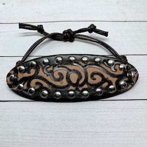 May include: A brown leather bracelet with a decorative design and silver studs. The bracelet has a tie closure.