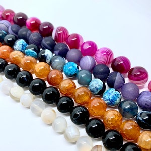 May include: Rows of gemstone beads in various colors, including pink, purple, blue, orange, black, and white. The beads have different textures, some smooth and others faceted. The beads are arranged diagonally against a white background, showcasing their diverse colors and patterns.