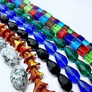 Op de afbeelding: Assortiment van kleurrijke glazen kralen, waaronder traanvormige kralen in zwart, blauw en groen, vierkante kralen in rood, blauw, groen en bruin, en oranje en gele bloemvormige kralen.