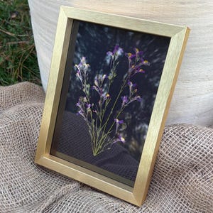 以下が含まれることがあります： 金色のフレームに入った長方形の絵で、植物の展示がされています。このアートワークは、暗い背景に繊細な紫と白の花が描かれています。フレームは角度がついており、質感のあるベージュ色の布の上に置かれています。